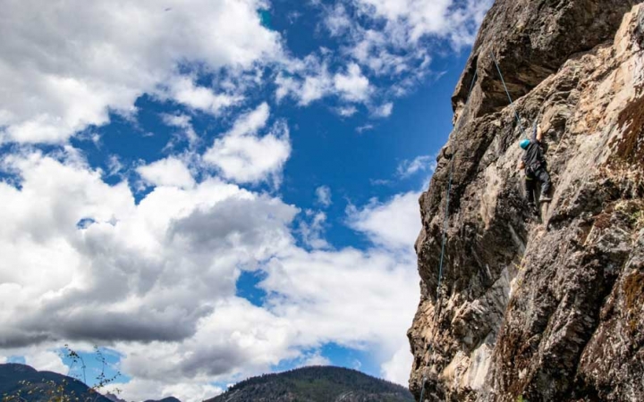 A person wearing safety gear is secured by ropes as they climb a tall rock wall. The sky is blue with white clouds.